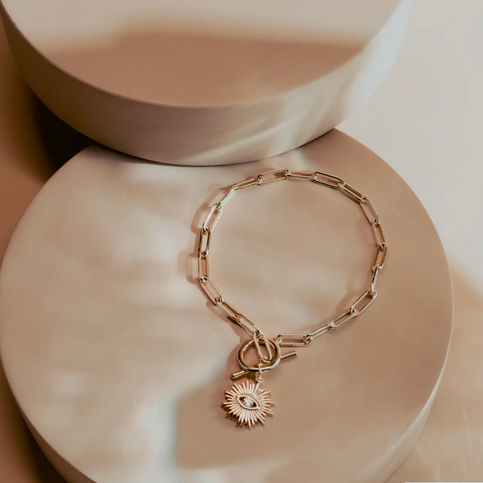 The Golden Eye Bracelet blends sleek design with symbolic detail. Crafted in 14k gold plated over silver, it features a bold paperclip chain and a front toggle clasp accented by a radiant sunburst charm set with a clear crystal eye. Polished and versatile, it brings a touch of strength and modern refinement to any style.