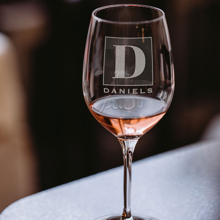 Stemmed wine glass on a table. The glass has an etched design centered. The design is of a square with swirly horizontal lines, and in the middle is the letter "D" in caps. Below the square is the name "DANIELS" in all caps.