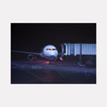 The artwork 'RED EYE' by Alex Devereux captures a commercial aircraft at night, illuminated by ambient terminal lighting. The composition shows a modern passenger jet connected to a jet bridge, with dramatic purple and blue tones creating a moody atmosphere. The aircraft's nose and cockpit windows glow against the dark background, emphasizing the nighttime airport setting, acrylic on canvas, 30x40 inches.