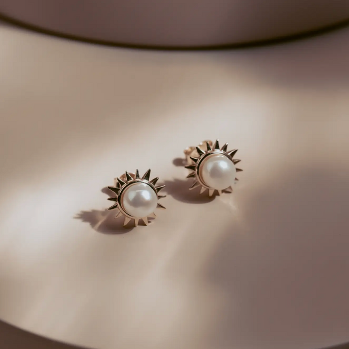 The Solar Pearl Studs capture the warmth of the sun in a timeless design. Each luminous freshwater pearl is framed by a burst of golden rays, crafted in 14k gold plate over sterling silver. Elegant yet bold, they bring effortless sophistication to any look—perfect for day or night.