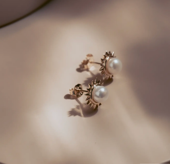 The Solar Pearl Studs capture the warmth of the sun in a timeless design. Each luminous freshwater pearl is framed by a burst of golden rays, crafted in 14k gold plate over sterling silver. Elegant yet bold, they bring effortless sophistication to any look—perfect for day or night.
