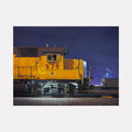 The artwork 'UNION-579' by Alex Devereux captures a striking yellow Union Pacific locomotive against a deep blue night sky. The train's number 579 is prominently displayed on its side, with industrial details and mechanical components visible beneath. The dramatic nighttime lighting creates strong contrasts, highlighting the train's vibrant color and metallic textures in this 12x16 inch acrylic painting on canvas.